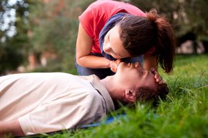 First aid - Look, listen and feel for breathing. Woman is performing first and on a man laying in the grass. She is pulling his head back, listening to his breath and looking at his chest to check if he is breathing. The man is laying unconscious.